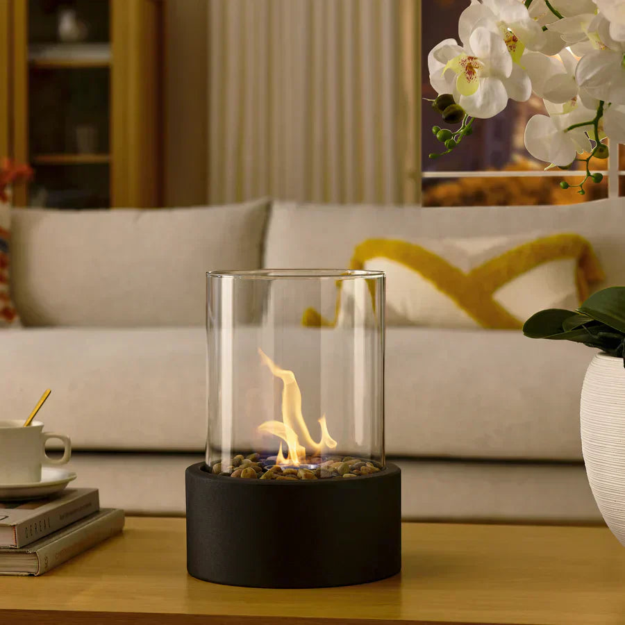 Modern tabletop fireplace with glass cylinder and black base on a wooden table, surrounded by books and a white orchid, in a cozy living room setting.