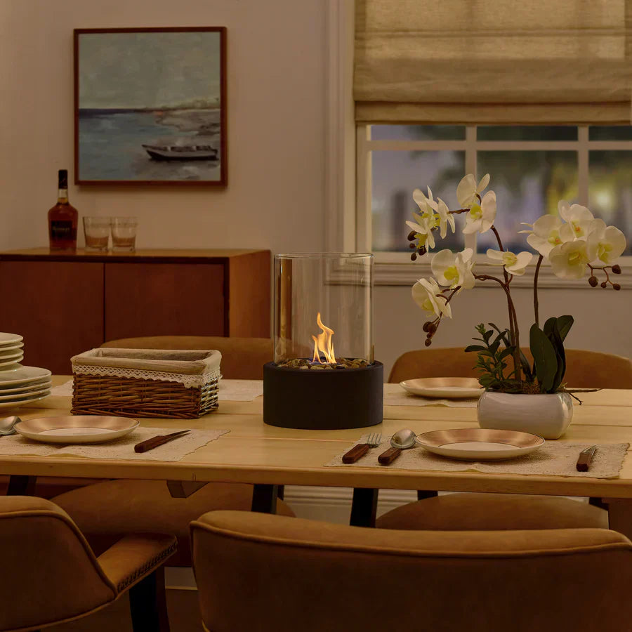 Tabletop fire pit on dining table with elegant orchid centerpiece, surrounded by plates and cutlery, in cozy dining room setting.