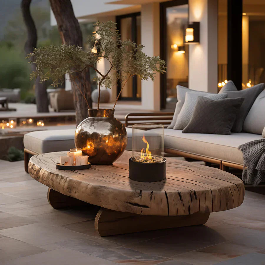 Rustic outdoor patio with a wooden coffee table featuring a modern tabletop fire pit, candles, and a decorative vase, surrounded by cozy seating.