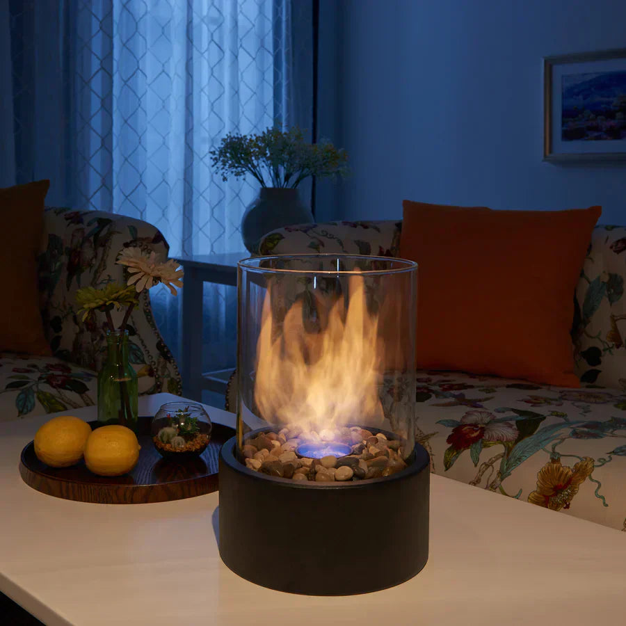 Tabletop fire pit with glass cylinder, black base, and decorative stones, set on a white table in a cozy living room. Perfect for indoor ambiance.