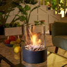 Tabletop fire pit with glass cylinder, decorative stones, and vibrant flame on a glass table. Indoor setting with plants and fruit bowl. Cozy ambiance.