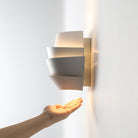 Modern white wall sconce with layered design, illuminating a hand. Contemporary lighting fixture, minimalist home decor, energy-efficient LED.