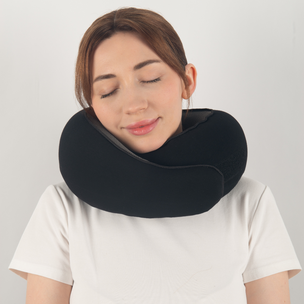 Woman wearing a black travel neck pillow, eyes closed, against a white background. Comfortable travel accessory, ergonomic neck support, relaxation.