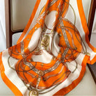 Luxury silk scarf with orange and white equestrian print, featuring intricate chain and belt design, draped over a chair. Fashion accessory.