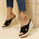 Women's black wedge sandals with braided platform, open toe design, paired with frayed jeans and heart anklet. Casual summer footwear.