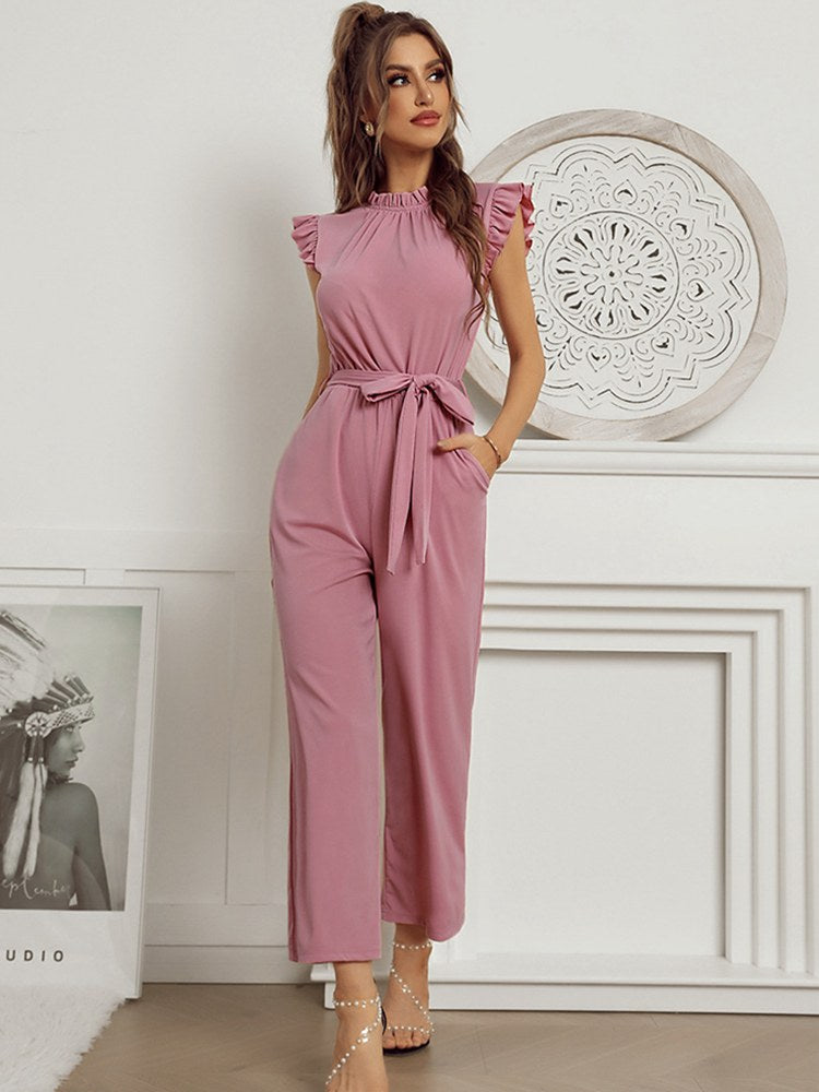 Woman in stylish pink ruffle-sleeve jumpsuit with tie waist, standing in elegant room with decorative wall art. Fashionable women's clothing.