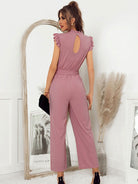 Woman in pink ruffled sleeveless jumpsuit, holding a black clutch, standing by an arched mirror with pampas grass decor, modern fashion style.