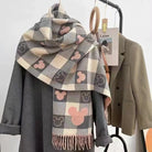 Plaid scarf with Mickey Mouse pattern draped over a gray coat on a hanger, featuring beige, pink, and gray tones. Fashion accessory display.