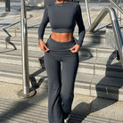 Woman in stylish dark gray workout set with long-sleeve crop top and high-waisted pants, standing on urban steps. Fitness fashion, activewear.