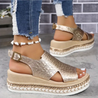 Gold platform sandals with textured straps and buckle, paired with distressed jeans and a shell anklet, against a brick wall background. Fashion footwear.