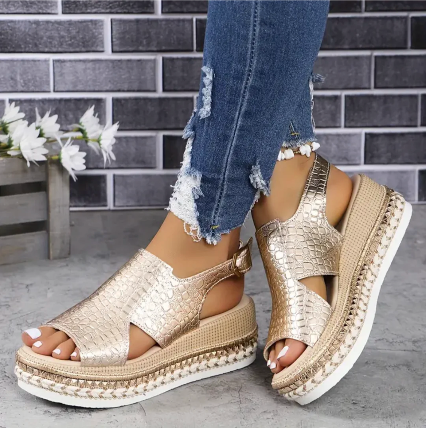 Gold platform sandals with textured design, paired with ripped blue jeans, against a brick wall background. Fashionable summer footwear for women.