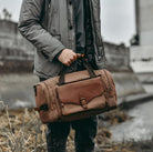 Man holding stylish brown leather duffel bag with shoulder strap, wearing a gray jacket. Perfect for travel, gym, or weekend trips.