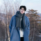 Young man in blue coat and gray scarf standing in snowy forest, winter fashion, outdoor portrait, serene landscape, stylish cold weather attire.