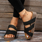 Women's black braided strap sandals with platform sole, worn with black leggings and anklet. Stylish summer footwear on wooden background.