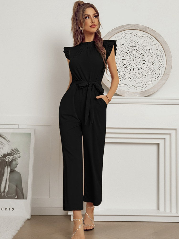 Woman in elegant black jumpsuit with ruffled sleeves and waist tie, standing in stylish room. Fashionable outfit, chic women's clothing, modern style.