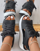 Black open-toe sneakers with white soles, featuring mesh design and laces, worn with gold anklets. Trendy women's footwear for casual style.