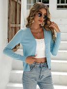 Woman in blue cardigan, white crop top, and jeans, wearing sunglasses, standing on white steps.