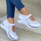 White mesh Mary Jane sneakers with gray accents, featuring a comfortable strap design, worn with blue jeans. Perfect for casual and sporty looks.