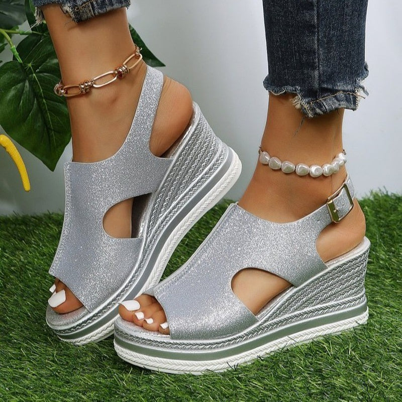 Silver wedge sandals with open toe design, ankle strap, and textured platform sole on grass. Fashionable women's footwear, summer style.