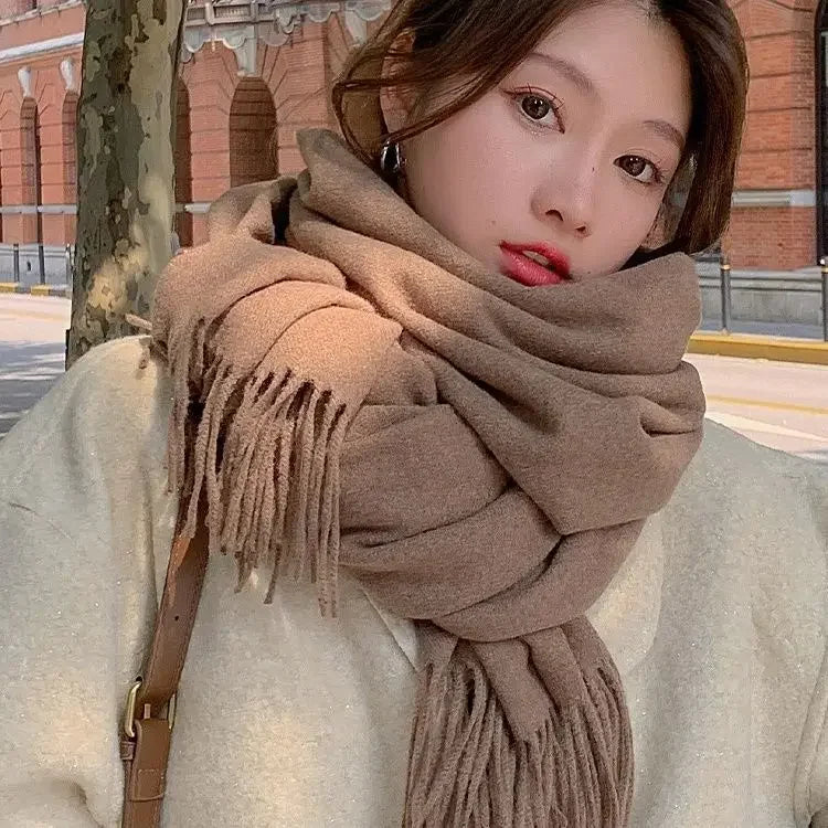 Woman wearing a large brown scarf and cream coat, standing outdoors with a brick building background. Fashionable winter outfit, cozy style.