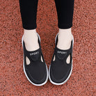 Black mesh slip-on sneakers with "SPORT" text, featuring cut-out design, worn on red textured surface. Comfortable, casual footwear for active lifestyle.