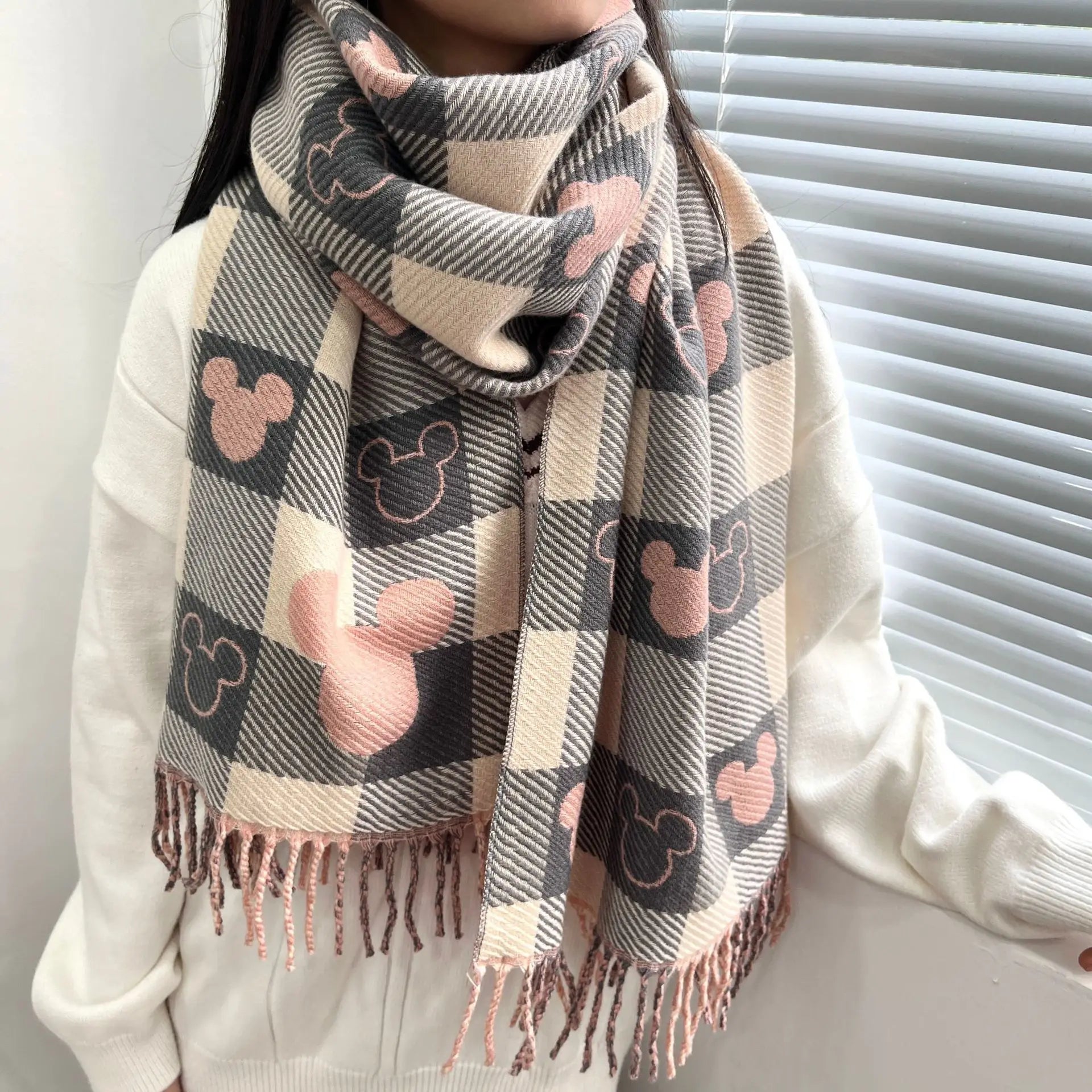 Plaid scarf with pink and gray Mickey Mouse pattern, worn by a person in a white sweater, featuring fringe details. Cozy winter fashion accessory.