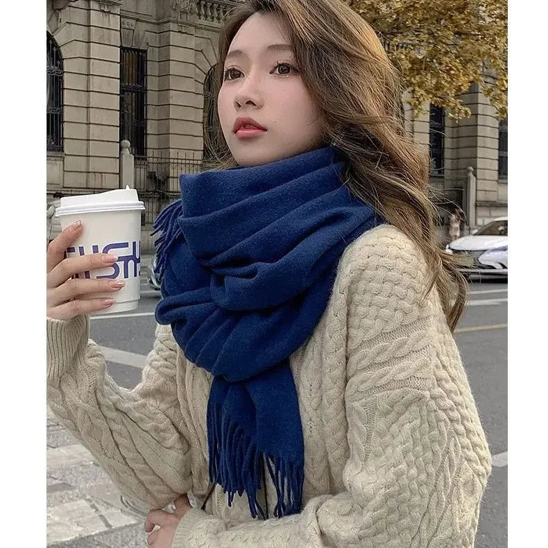 Woman in cozy cream cable-knit sweater and blue scarf holding a coffee cup outdoors. Fashionable fall outfit, urban street style, autumn fashion.