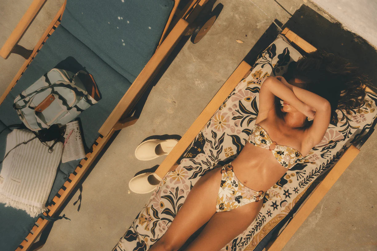 Woman sunbathing in floral bikini on patterned lounge chair, next to a blue cushioned chair with a striped bag and sandals. Summer relaxation scene.