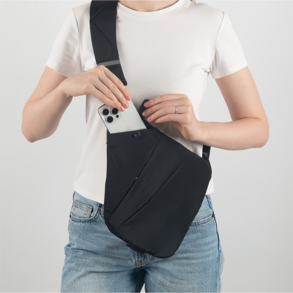 Woman placing smartphone in black crossbody sling bag, wearing white t-shirt and jeans. Stylish, functional accessory for secure, hands-free convenience.