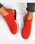 Red slip-on sneakers with knit upper, white sole, and black tread, worn with frayed hem jeans on marble floor. Stylish, comfortable footwear.