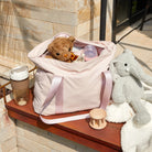 Pink diaper bag with teddy bear, baby bottle, and toys on a patio ledge. Includes a reusable coffee cup and plush elephant. Perfect for parents on-the-go.