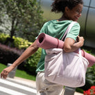 Woman carrying pink yoga mat and tote bag, wearing green shirt and beige pants, walking outdoors. Fitness lifestyle, activewear, exercise gear.