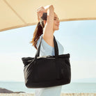 Woman in blue dress holding a large black tote bag outdoors under a canopy. Stylish summer fashion, beach accessory, casual chic look.