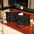 Two black tote bags on a wooden bench, featuring sturdy handles and spacious design. Nearby are a wallet, a tumbler, and a small retro radio.