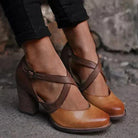 Women's brown leather block heel shoes with cross-strap design, worn with black pants. Stylish, comfortable footwear for casual or formal occasions.