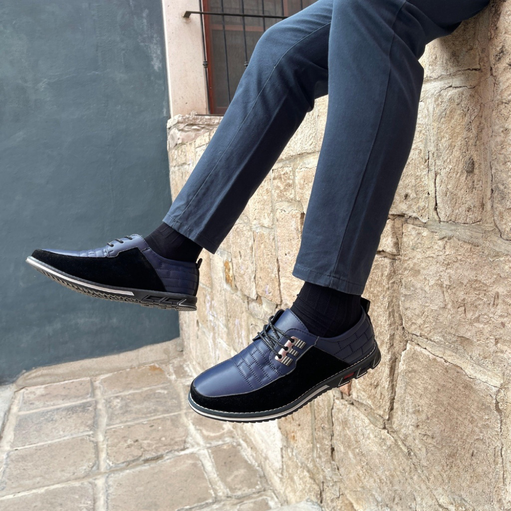 Men's stylish navy blue leather shoes with black accents, worn with dark pants, against a rustic stone wall. Fashionable footwear for casual or formal wear.