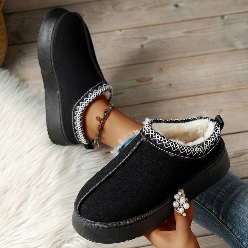 Black fleece-lined winter slippers with white stitching, worn by a person on a wooden floor. Cozy indoor footwear, perfect for cold weather comfort.