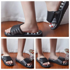 Black acupressure massage slippers with foot reflexology beads, worn by a person on a wooden floor. Comfortable therapeutic footwear.