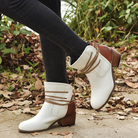 White leather ankle boots with brown heels and wraparound laces, worn with black jeans, on a leafy outdoor path. Fashionable fall footwear.