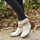 White leather ankle boots with brown accents and block heels, worn with black jeans, surrounded by autumn leaves. Fashionable fall footwear.