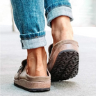 Casual brown suede slip-on shoes with textured rubber soles, worn with rolled-up blue jeans, walking on a concrete surface.