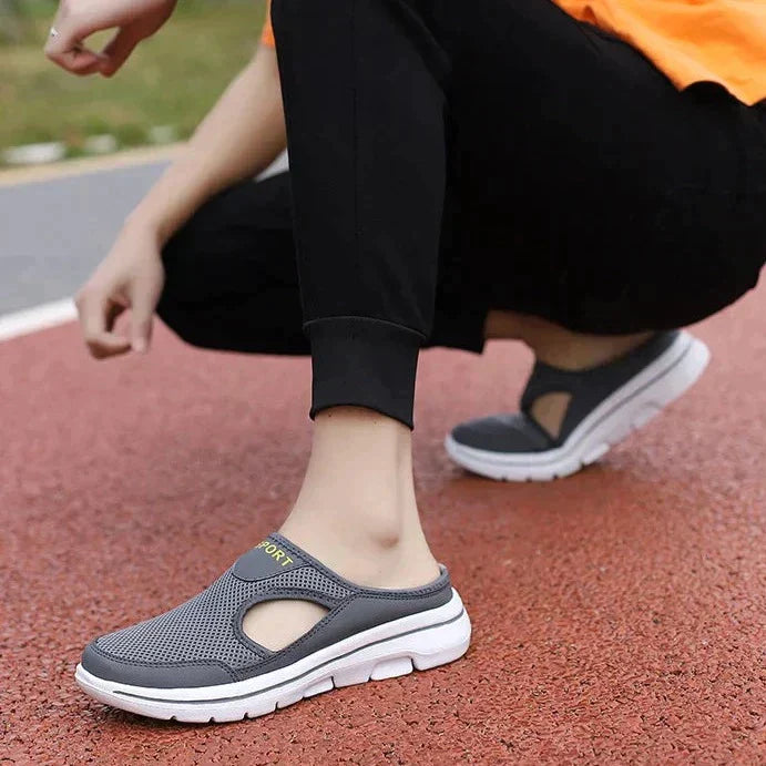 Gray slip-on athletic shoes with breathable mesh design and white soles, worn by a person in black pants on a red track. Comfortable walking footwear.