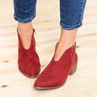 Red suede V-cut ankle boots with low block heels, worn with rolled-up blue jeans on a light wooden floor. Stylish women's fall footwear.