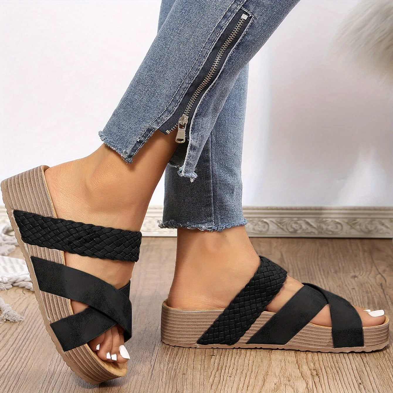 Black woven strap wedge sandals on a woman's feet, paired with blue jeans featuring a zipper detail, set against a neutral background.