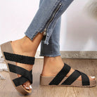 Black woven strap wedge sandals on a woman's feet, paired with blue jeans featuring a zipper detail, set against a neutral background.