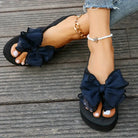 Black bow sandals on woman's feet with white pedicure, wearing anklets, on wooden floor. Fashionable summer footwear, casual style.