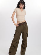 Woman wearing beige crop top and olive cargo pants, accessorized with a brown belt, standing against a white background. Fashionable casual outfit.