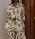 Woman wearing a stylish beige cactus print matching set, featuring a button-up shirt and pants, standing against a wooden door. Fashionable casual outfit.