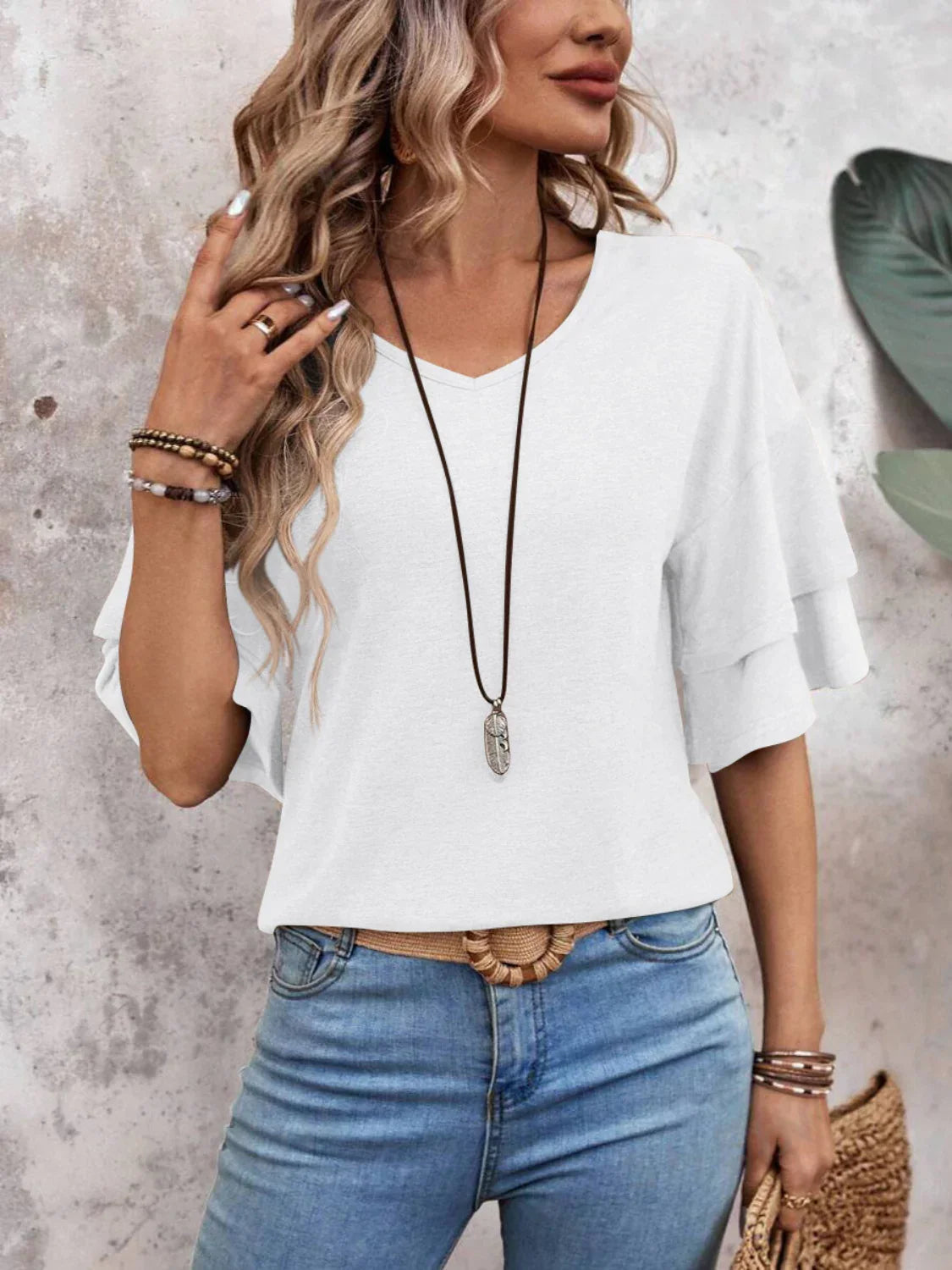 Woman in casual white ruffle sleeve top and blue jeans, accessorized with a long necklace and bracelets, standing against a textured wall.