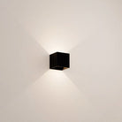 Modern black cube wall sconce with minimalist design, casting ambient light on white wall. Contemporary lighting fixture for home decor.